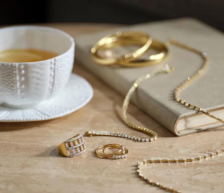 An assortment of yellow gold lab-grown diamond jewelry on a table with a book and coffee mug in frame. Shop lab grown diamond jewelry.